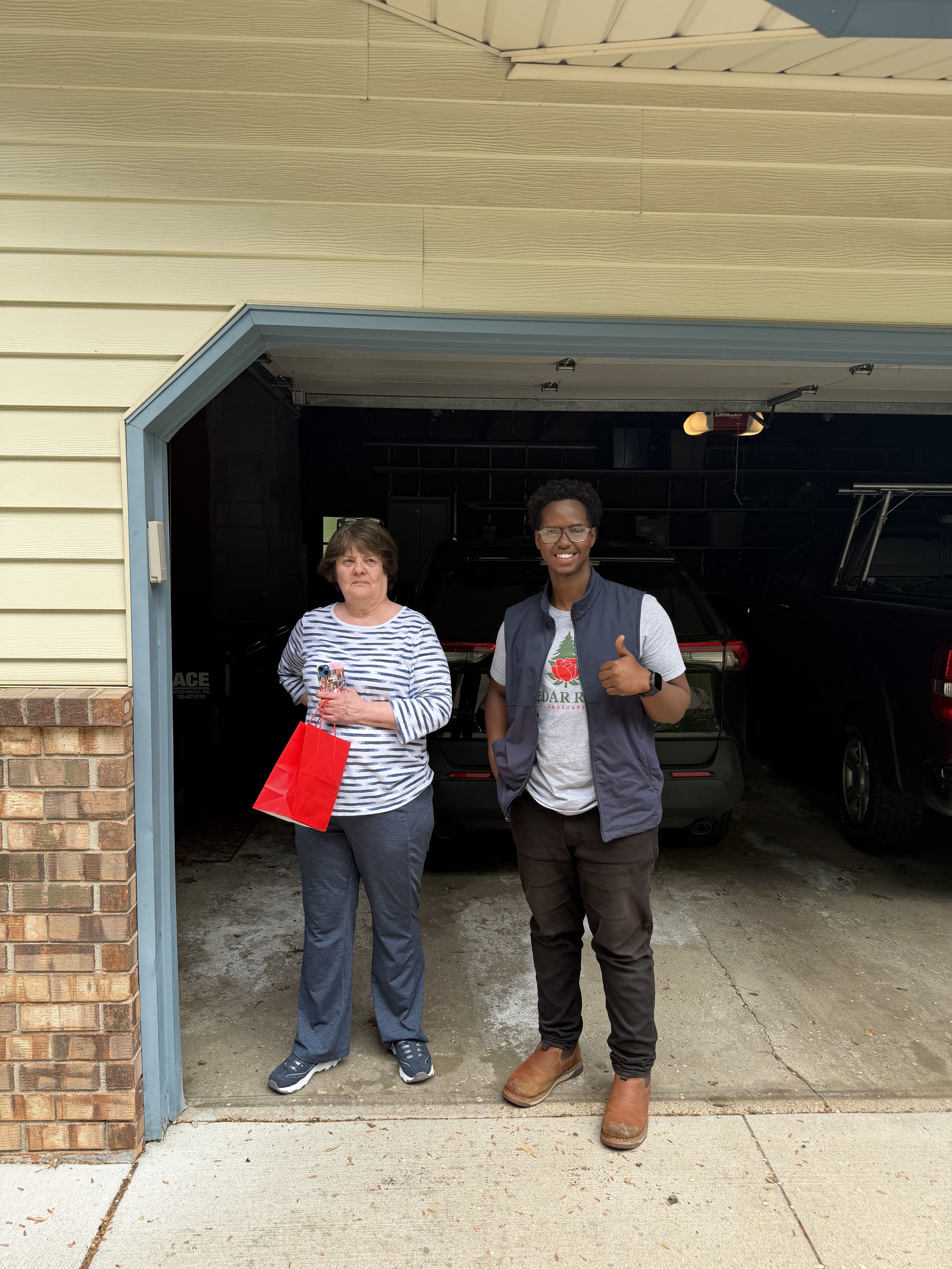 Woman and young man in garage, smiling, with a car in the background and a red bag.