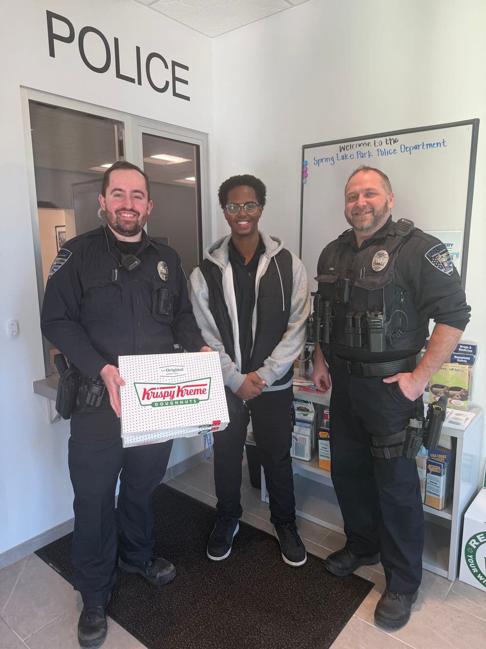 Three individuals at a police station, two officers and a young man holding a Krispy Kreme box.